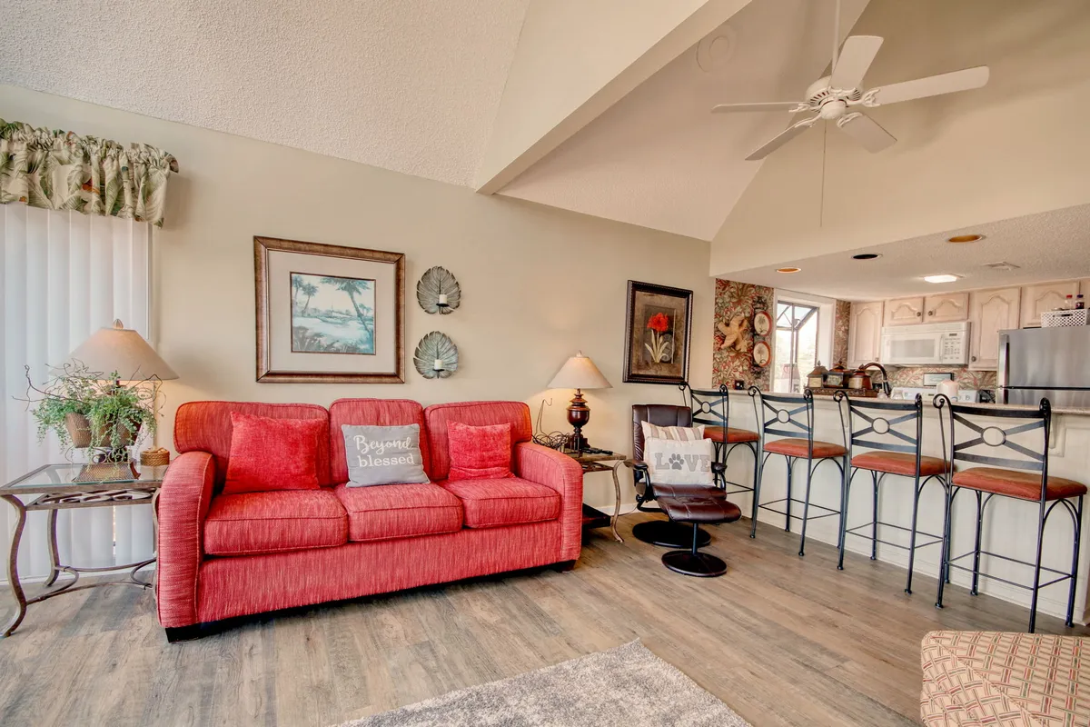 The image showcases a bright, inviting condo living space with a plush red couch, modern bar stools lined up at a breakfast bar, decorative wall art, and a warm floor lamp. The arrangement suggests a place where family and friends can relax, share meals, and create lasting memories together after a day at the beach.