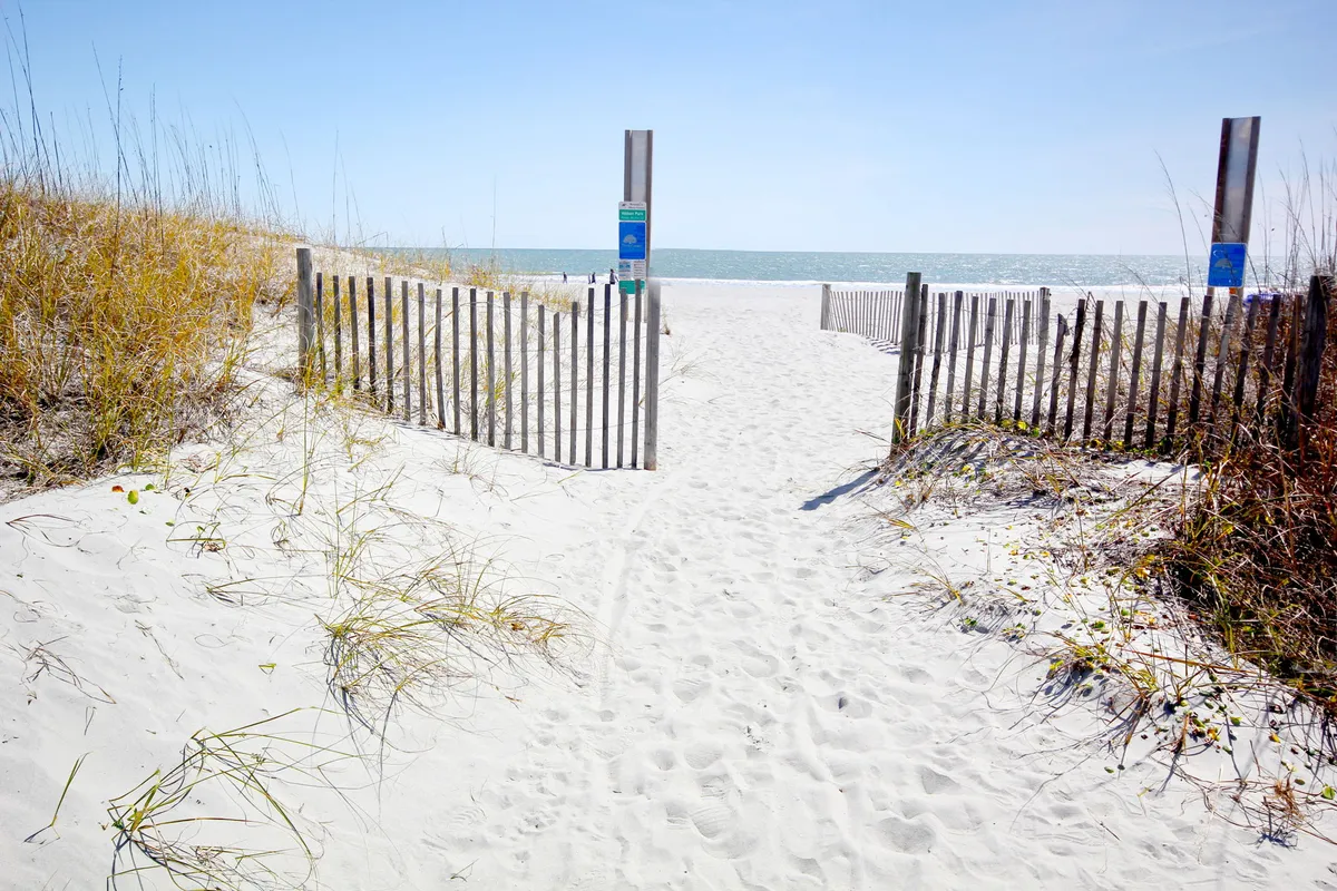 This image captures a wooden fence and pathway leading directly to the ocean, with beach grass and waves visible in the background. The scene showcases the ease of beach access and the inviting coastal environment awaiting just steps away.