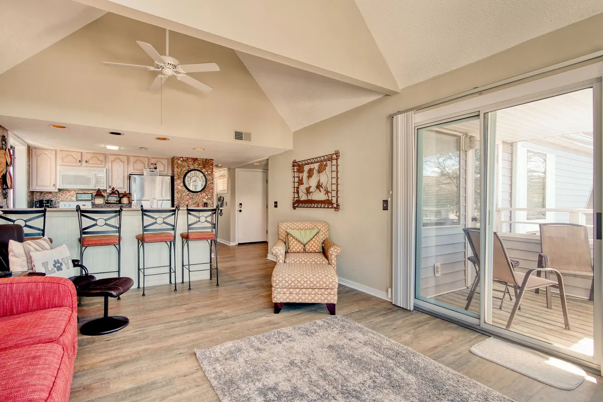 This scene captures a welcoming open-concept space combining the living room and part of the kitchen area. It includes a comfortable red sofa, bar stools at a kitchen counter, a charming armchair, and a large sliding glass door that allows natural light to flood the room. The space is arranged for easy conversation and shared activities.