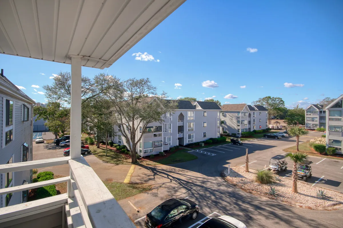 A serene balcony scene overlooking a lush courtyard with palm trees, a tree in view, and the golden glow of sunset visible. The setting offers a tranquil spot for relaxing or sharing a romantic evening, with the backdrop of the surrounding community buildings and peaceful outdoor ambiance.