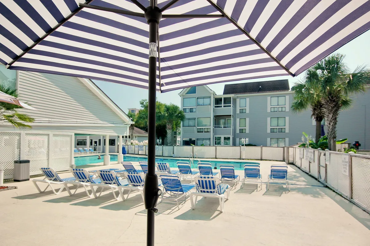 A sunlit swimming pool area surrounded by striped umbrellas, lounge chairs, palm trees, and apartment buildings. The pool is the focal point, with spaces for both sunbathing and swimming, suggesting a lively but relaxing atmosphere for families.