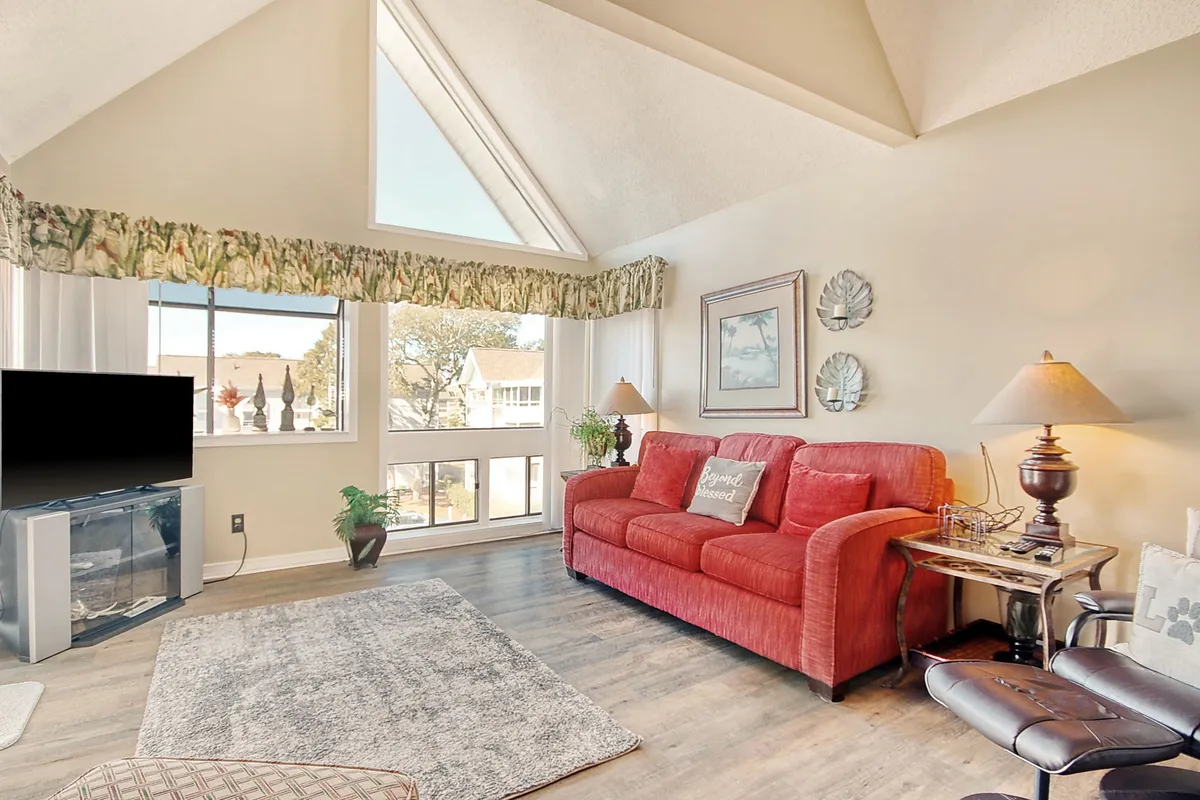 This image showcases a cozy living room area featuring a plush red sofa, expansive windows that let in ample natural light, a television, decorative wall art, and a table lamp. The sunlight creates a warm and welcoming environment ideal for relaxing, socializing, or unwinding after a busy day.