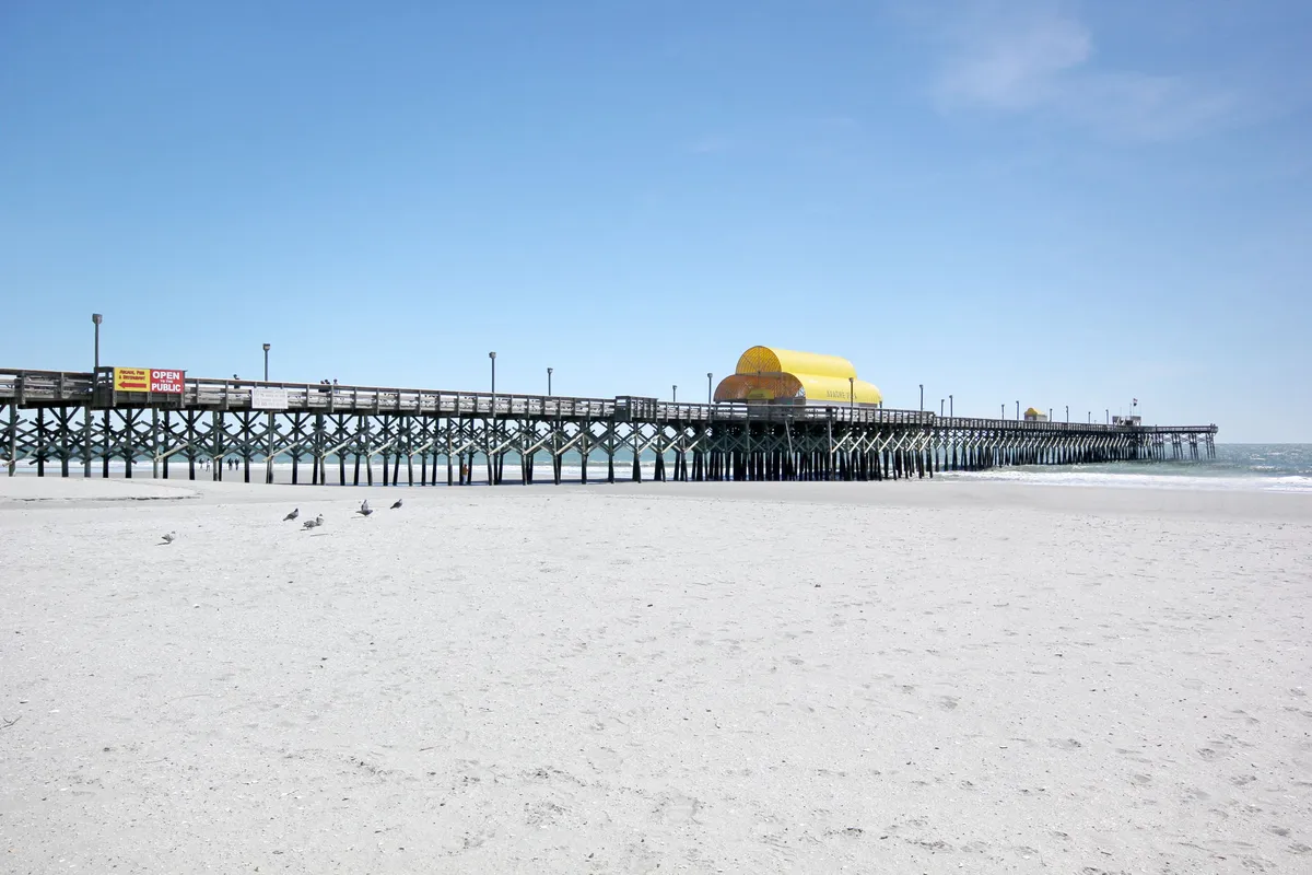 An iconic Myrtle Beach pier stretches into the ocean under a yellow canopy, with sand and gentle waves creating a classic coastal scene. The area is perfect for strolls, beachcombing, and enjoying the natural beauty of the beach at sunrise or sunset.