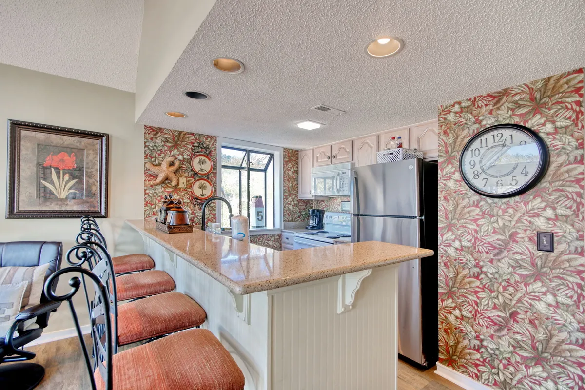 This view highlights a granite countertop, tropical wallpaper, a row of bar stools, and a large wall clock. The setup is ideal for casual dining, quick snacks, or socializing while cooking.