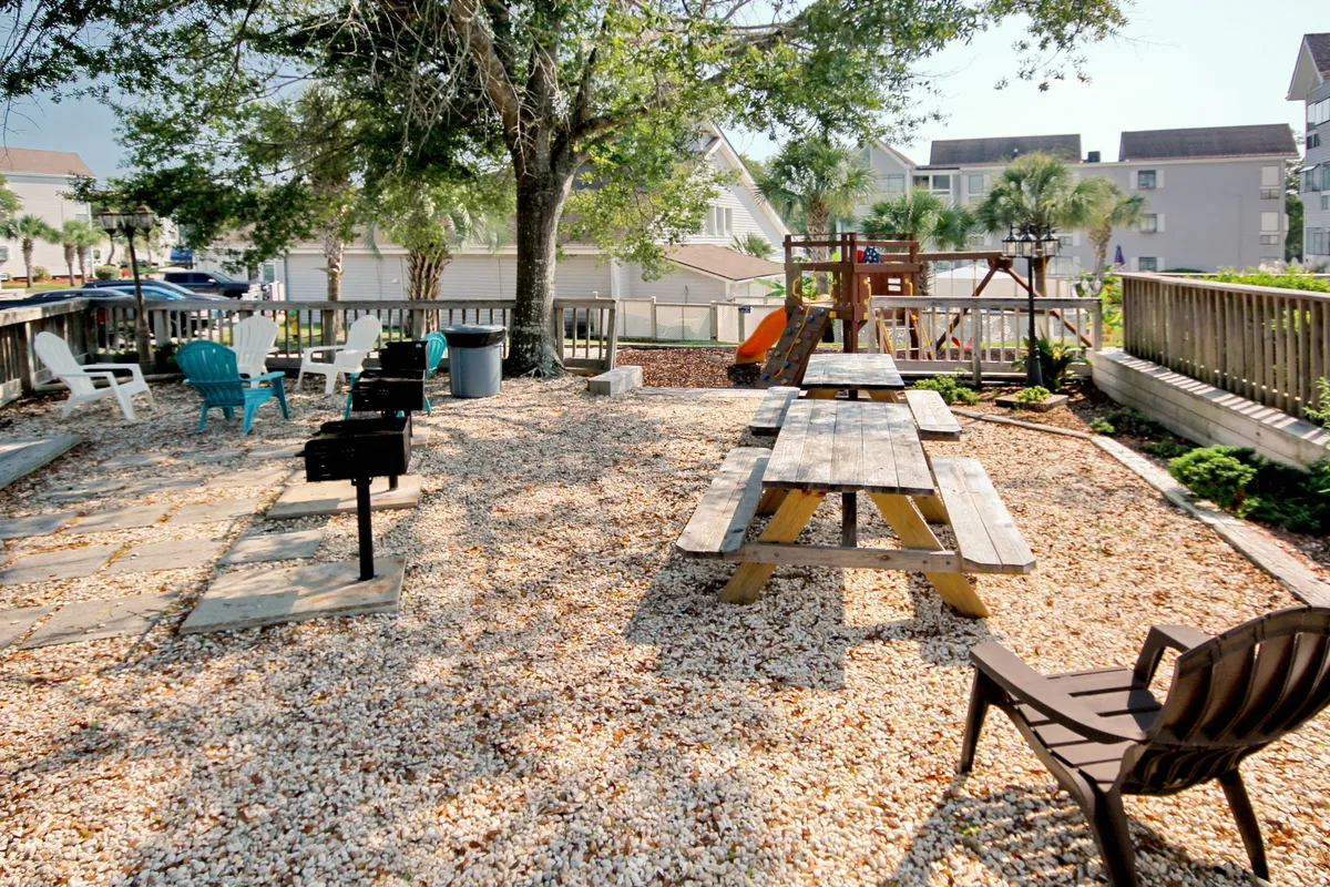 A lively outdoor area featuring picnic tables, a grill station, playset for children, and colorful seating all in a cheerful, family-friendly setting. The image captures the sense of togetherness and activity that the vacation community offers.