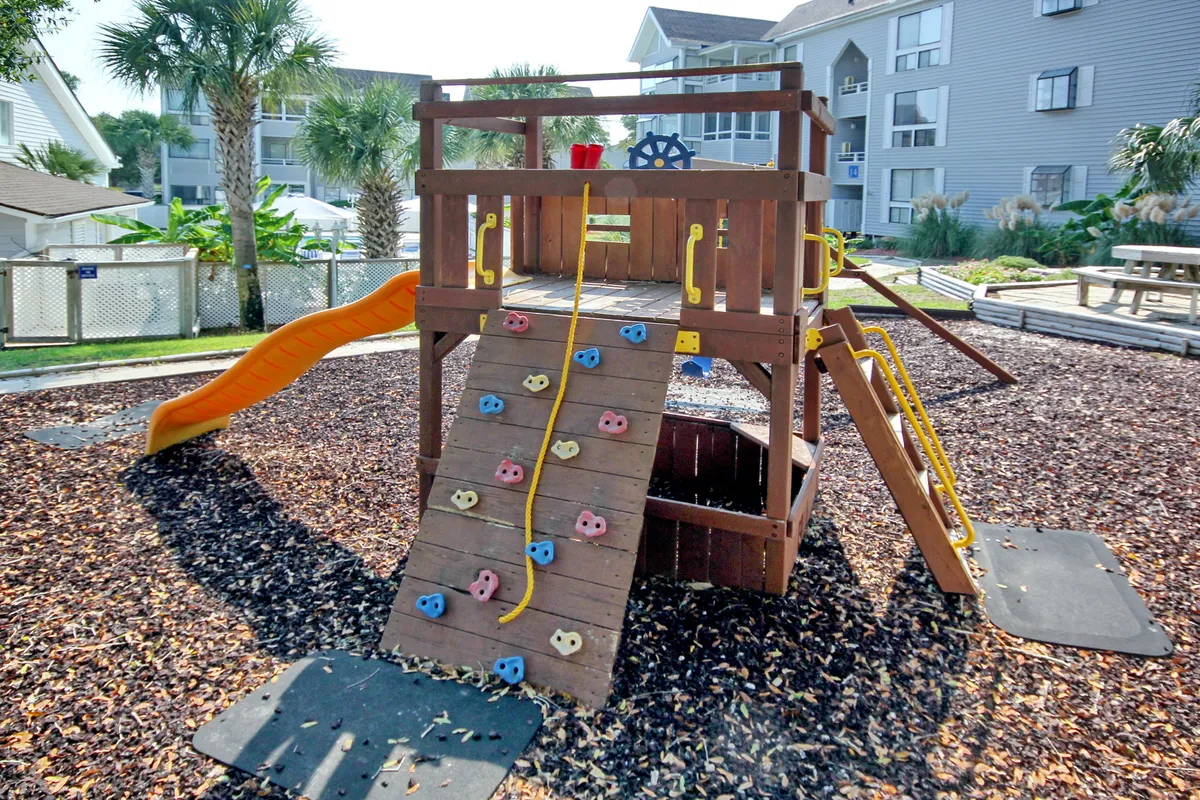 A colorful playground structure equipped with a slide and climbing wall, set in an outdoor community area. Children are seen playing and engaged in imaginative activities, surrounded by greenery and open space.