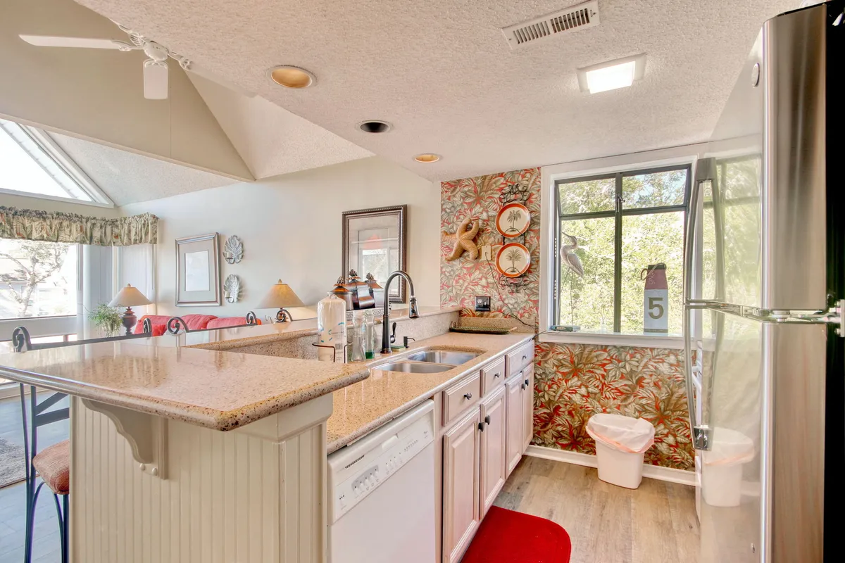 This image displays a vibrant kitchen scene featuring a central island with a granite countertop, colorful wallpaper, a window letting in ample natural light, and visible full-sized appliances such as a fridge. The setting is bright and inviting, with ample space for cooking and gathering.
