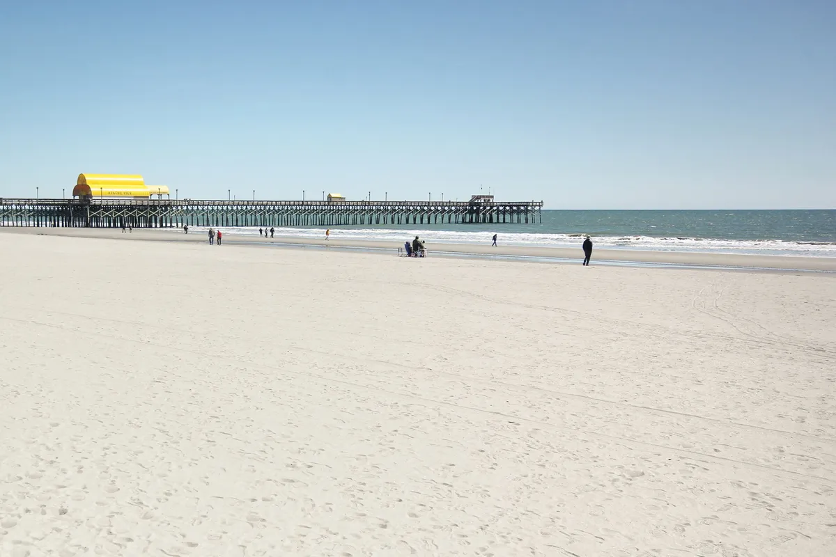 A lively beachfront scene with a pier stretching into the ocean, people strolling along the sandy shore, and a distinctive yellow building on the pier. The atmosphere is joyful and active, inviting extended outdoor enjoyment.