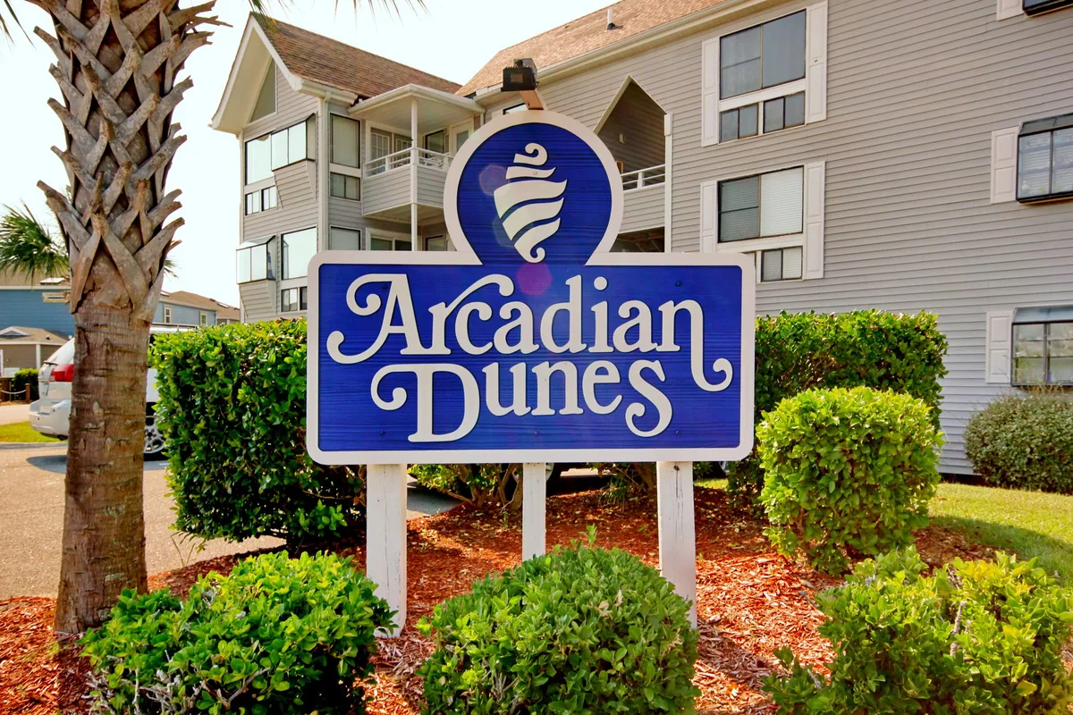 The entrance to Arcadian Dunes, featuring a prominent community sign surrounded by lush palm trees and well-maintained landscaping. The bright and cheerful scene immediately communicates a sense of welcome and belonging.